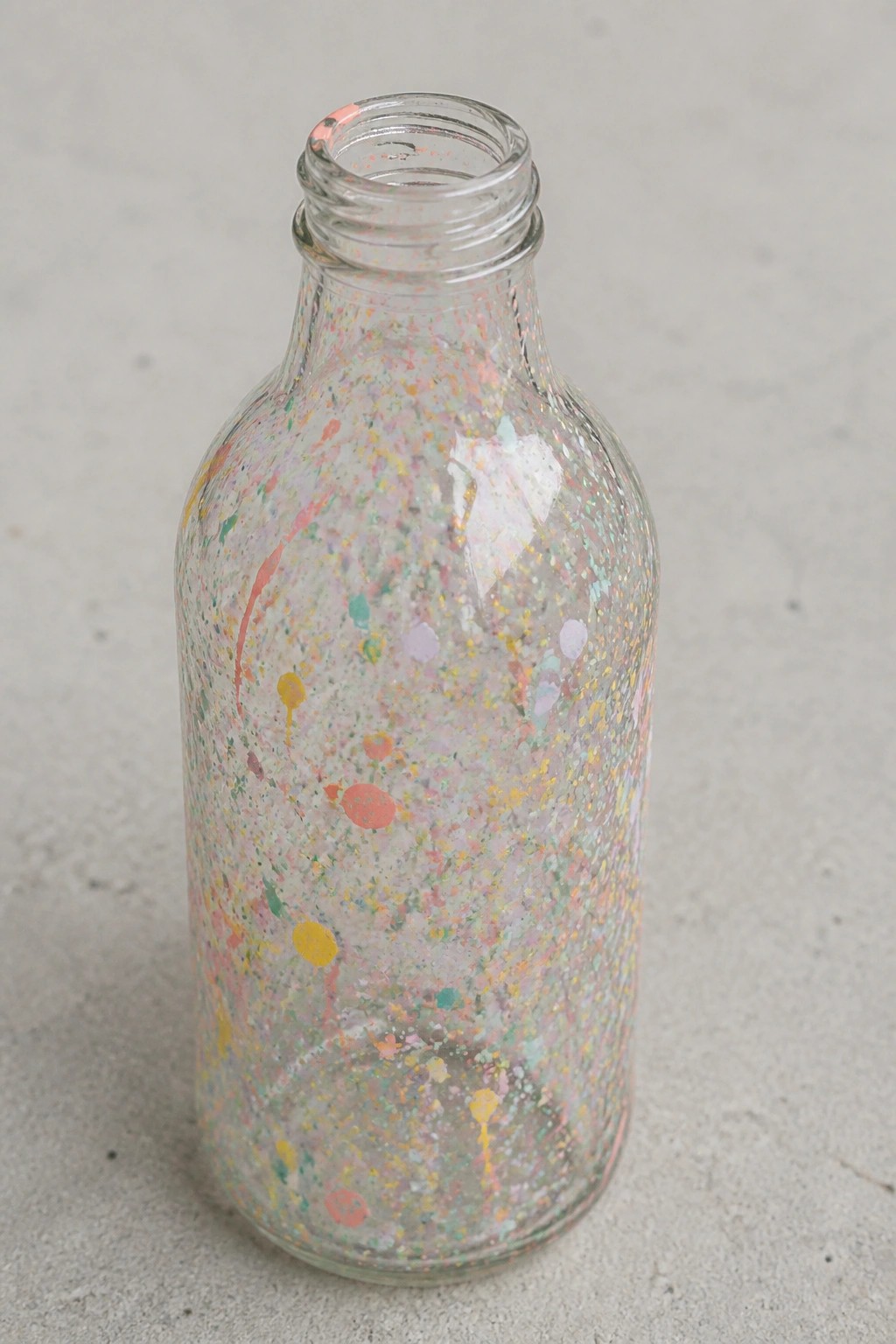 Clear glass bottle covered in vibrant multicolored paint splatters on gray surface
