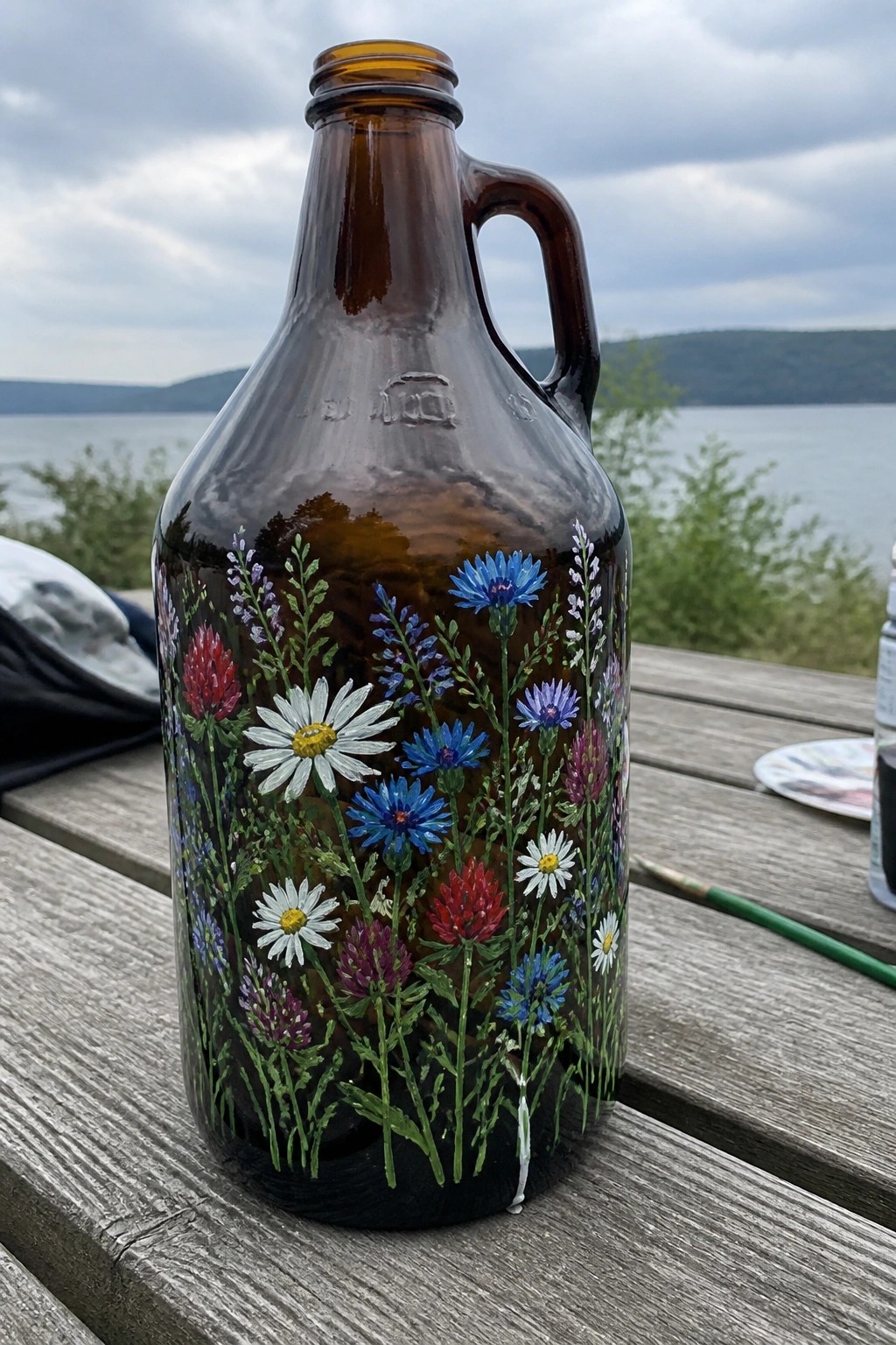 Brown glass jug with colorful painted wildflowers on wooden table by lake