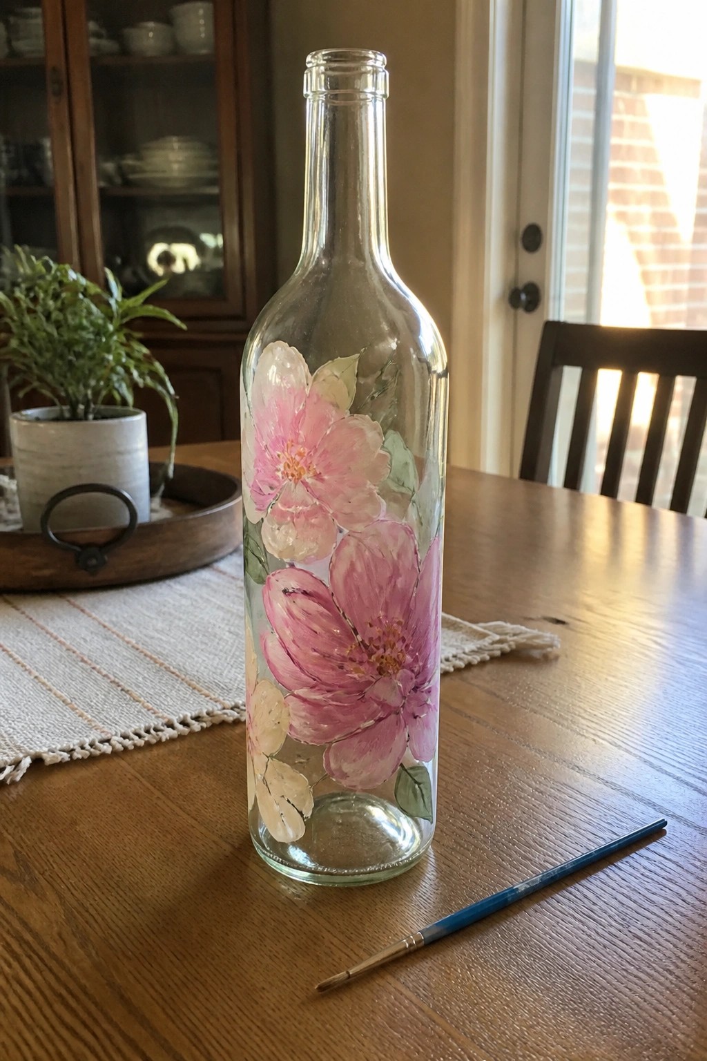 A clear glass bottle painted with large pink flowers sits on a wooden table next to a paintbrush.