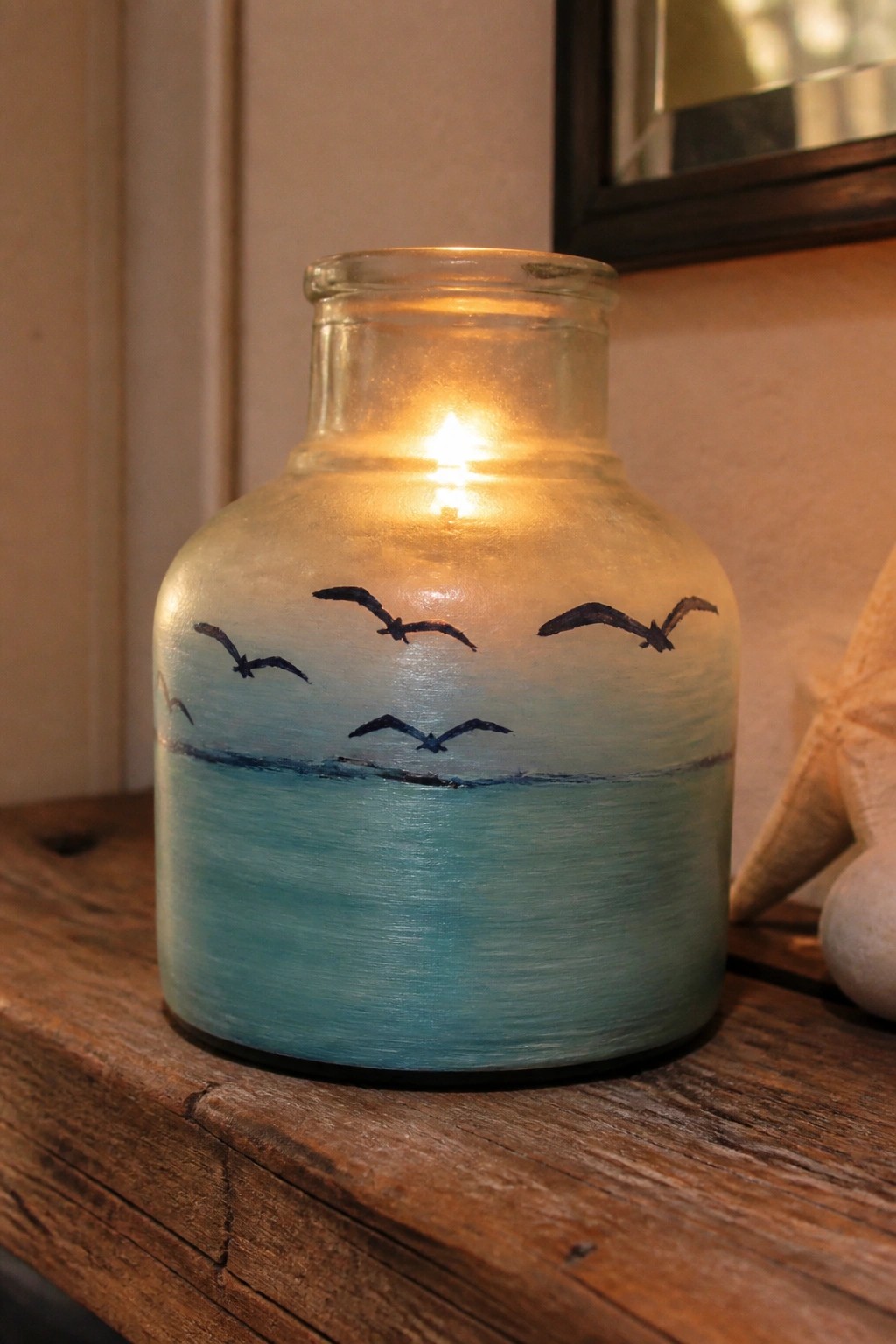 Lit candle glows inside glass jar painted with blue ocean and black birds.