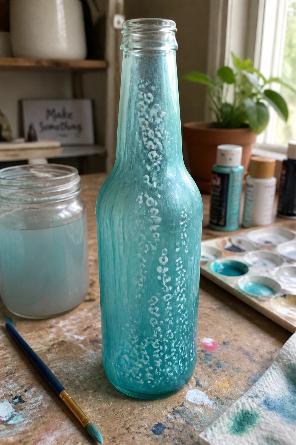 Turquoise painted glass bottle with white speckles amid art supplies on wooden table.