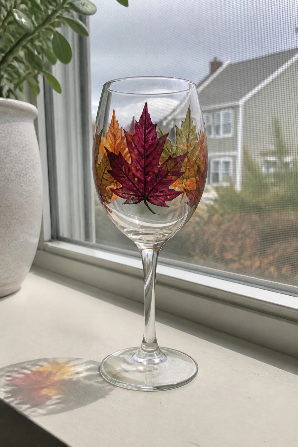 Wine glass with colorful autumn maple leaves on sunny windowsill with shadow