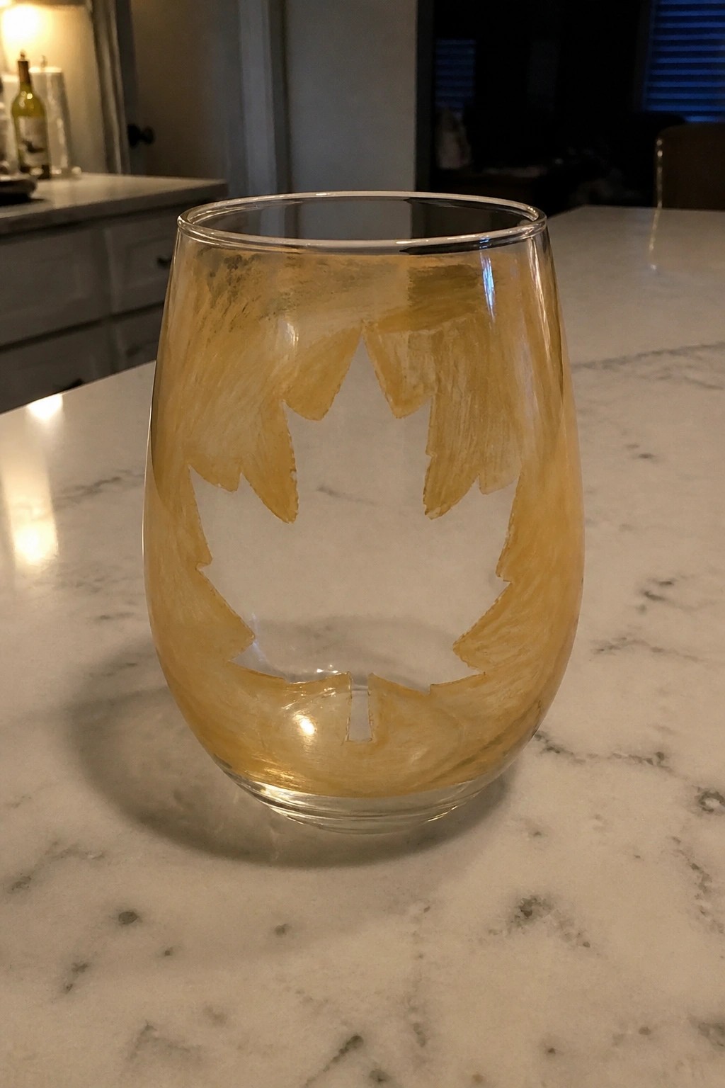 Stemless wine glass with gold maple leaf design on white marble countertop.
