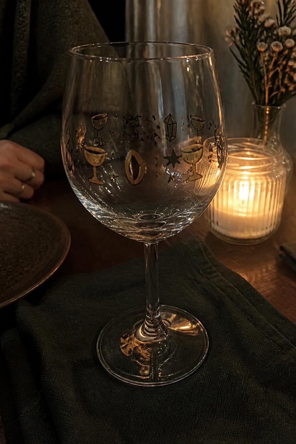 Wine glass etched with golden goblets, moon, stars on dark green cloth near candle