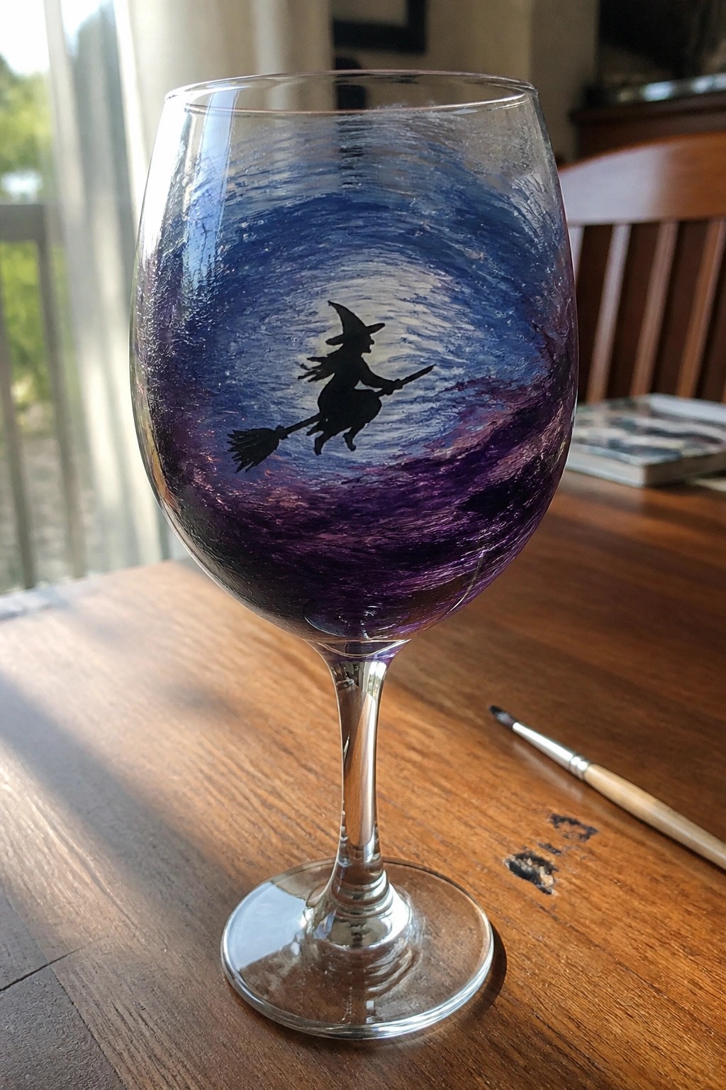Wine glass with black witch silhouette on broom amid swirling blue-purple sky paint.