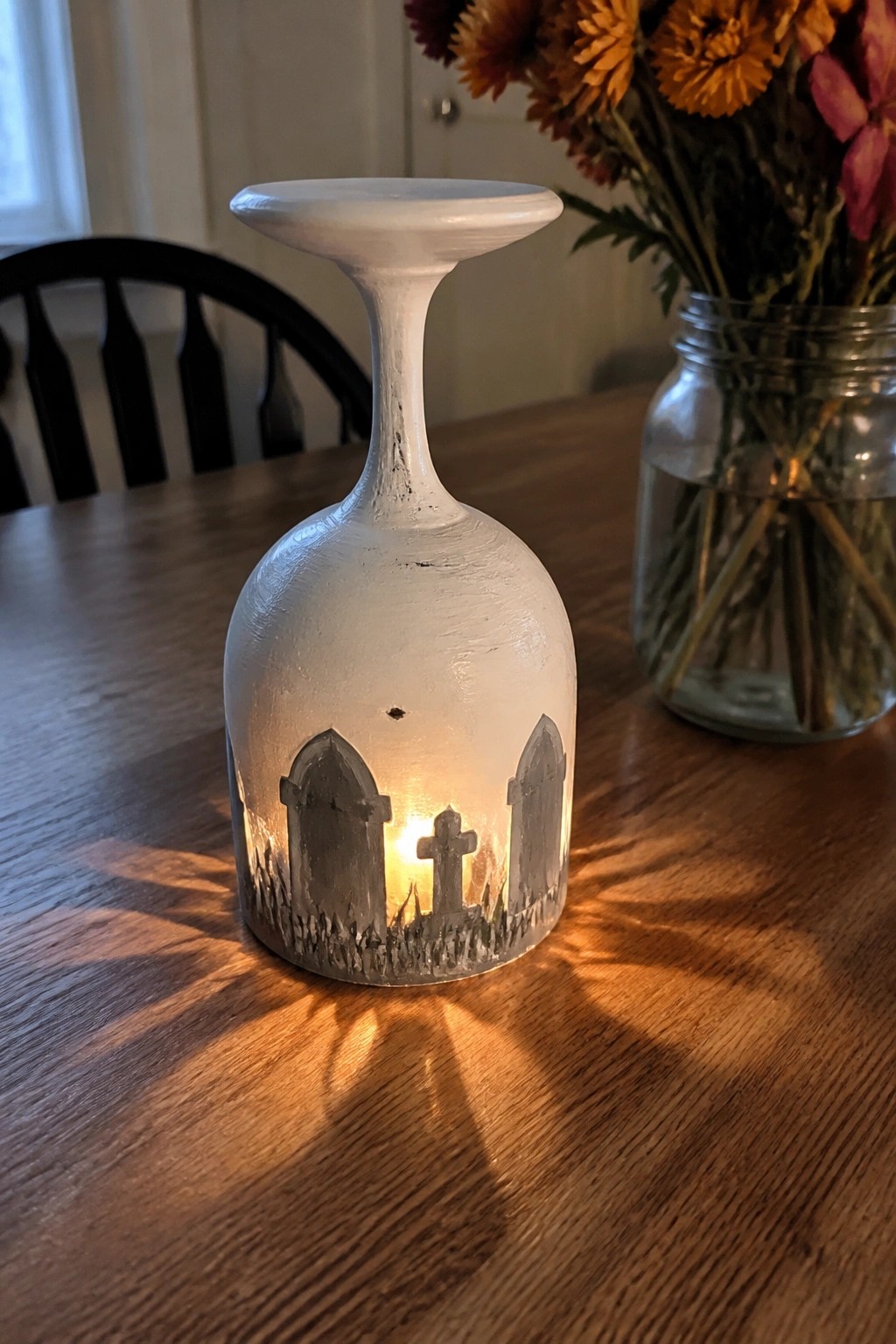 Lit white wine glass with glowing graveyard silhouettes on wooden table beside flowers