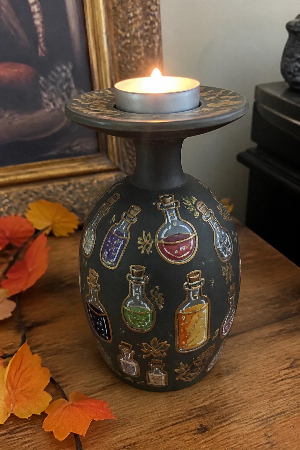 Dark vase painted with colorful potion bottles holds lit tealight amid autumn leaves.