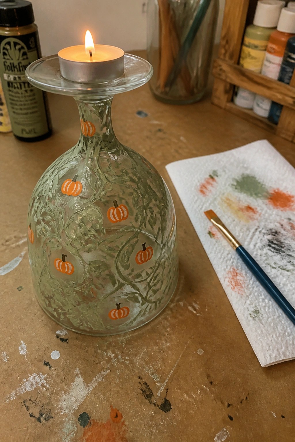 Glass bottle painted with green vines and orange pumpkins, holding lit tea light candle.