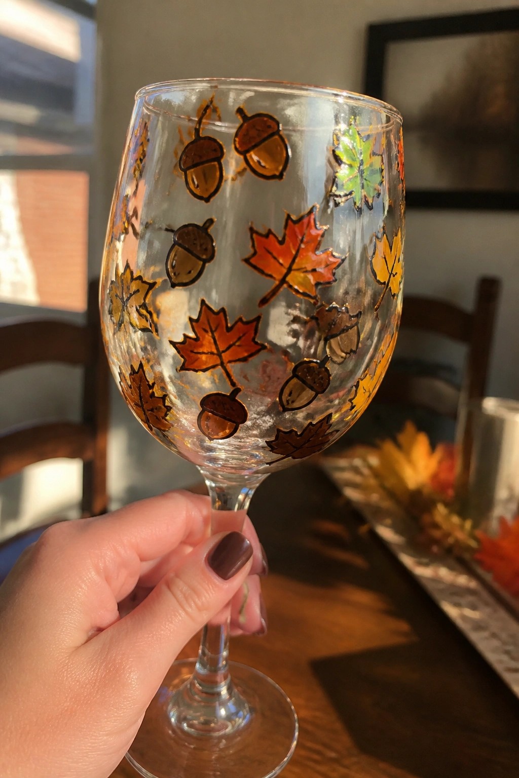 Hand holding wine glass painted with acorns and colorful autumn leaves.