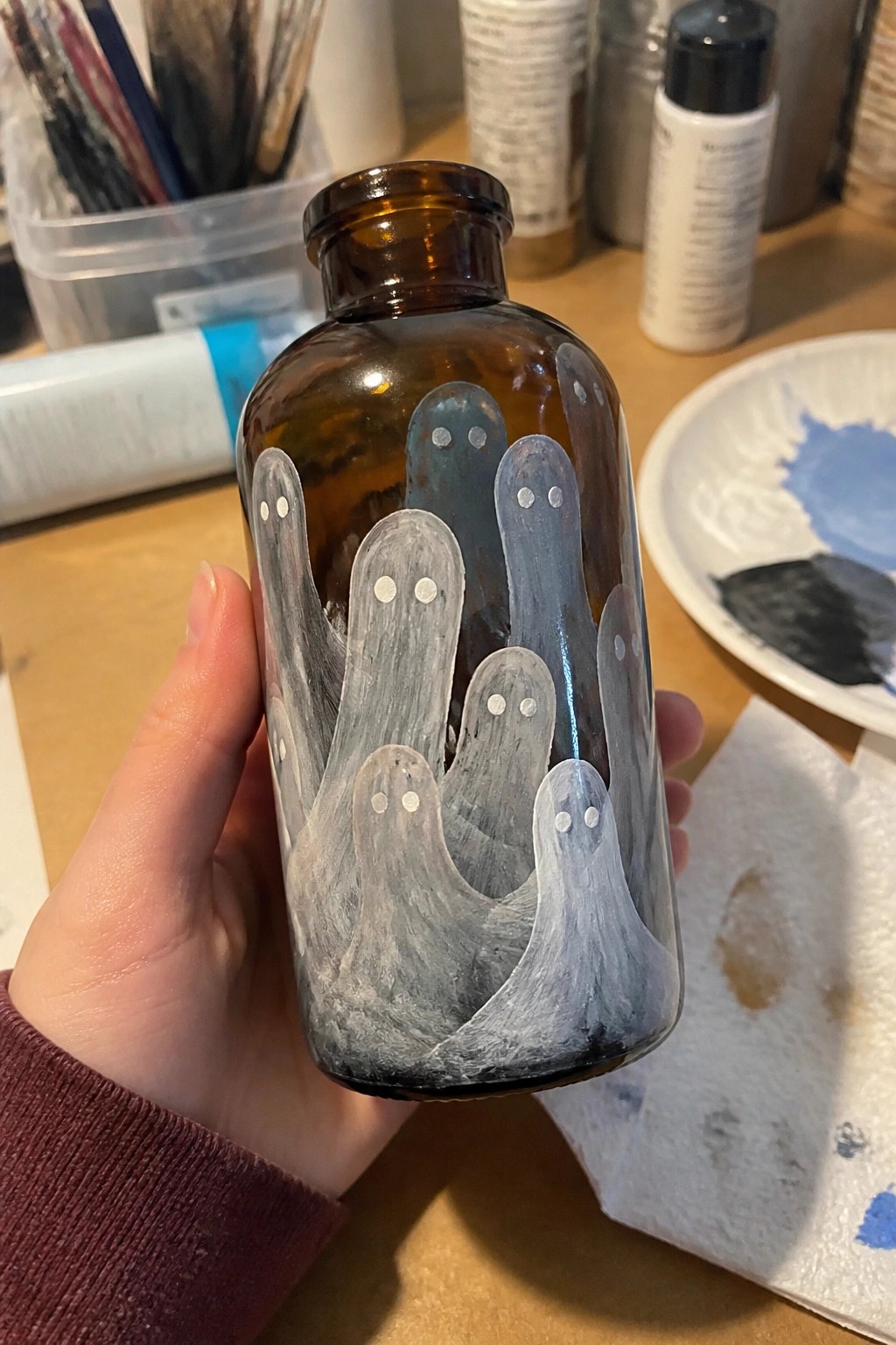 Hand holding brown bottle painted with white, gray and blue ghosts.