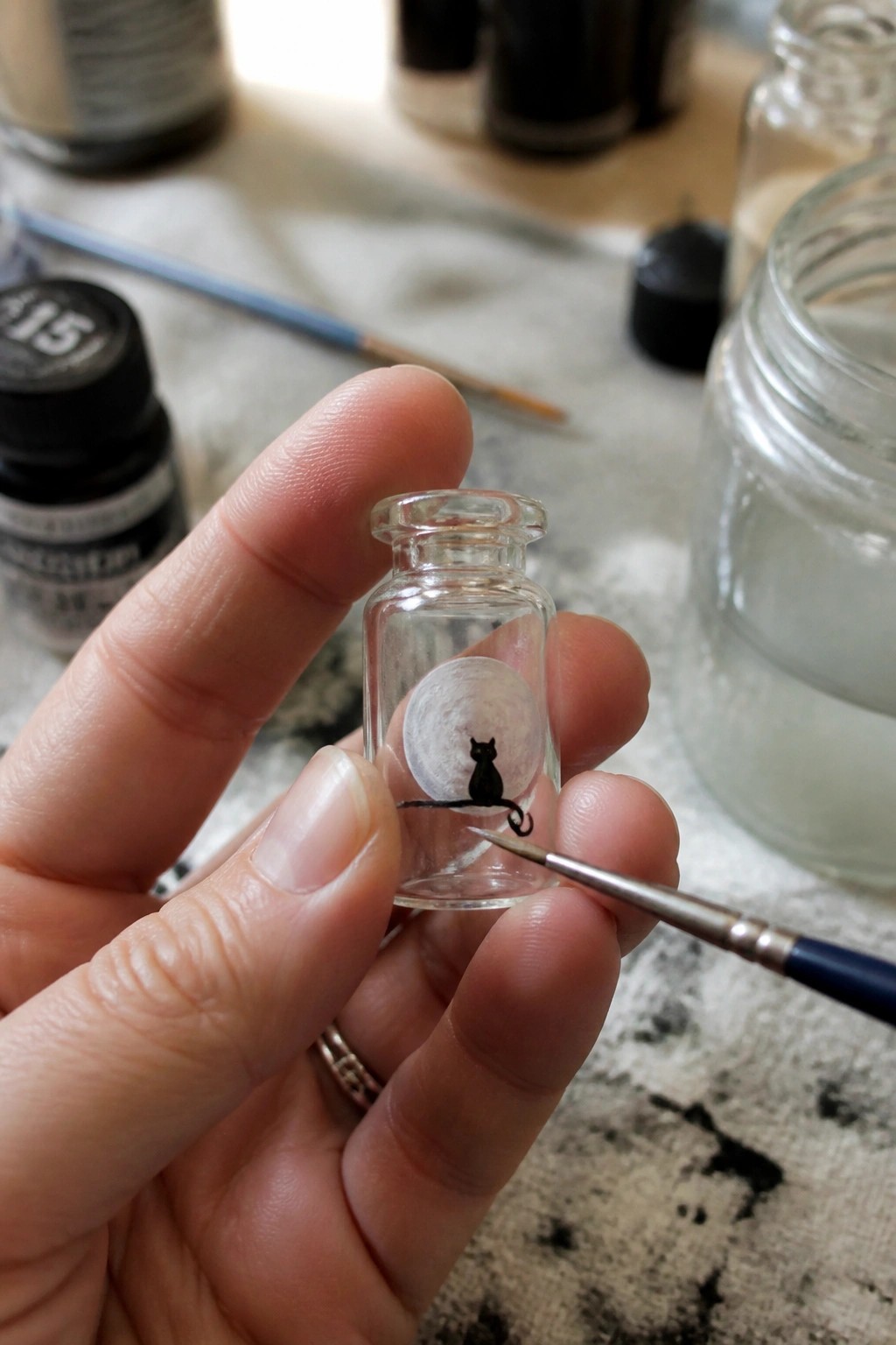 Hand holding tiny glass bottle with black cat silhouette before painted moon, brush nearby
