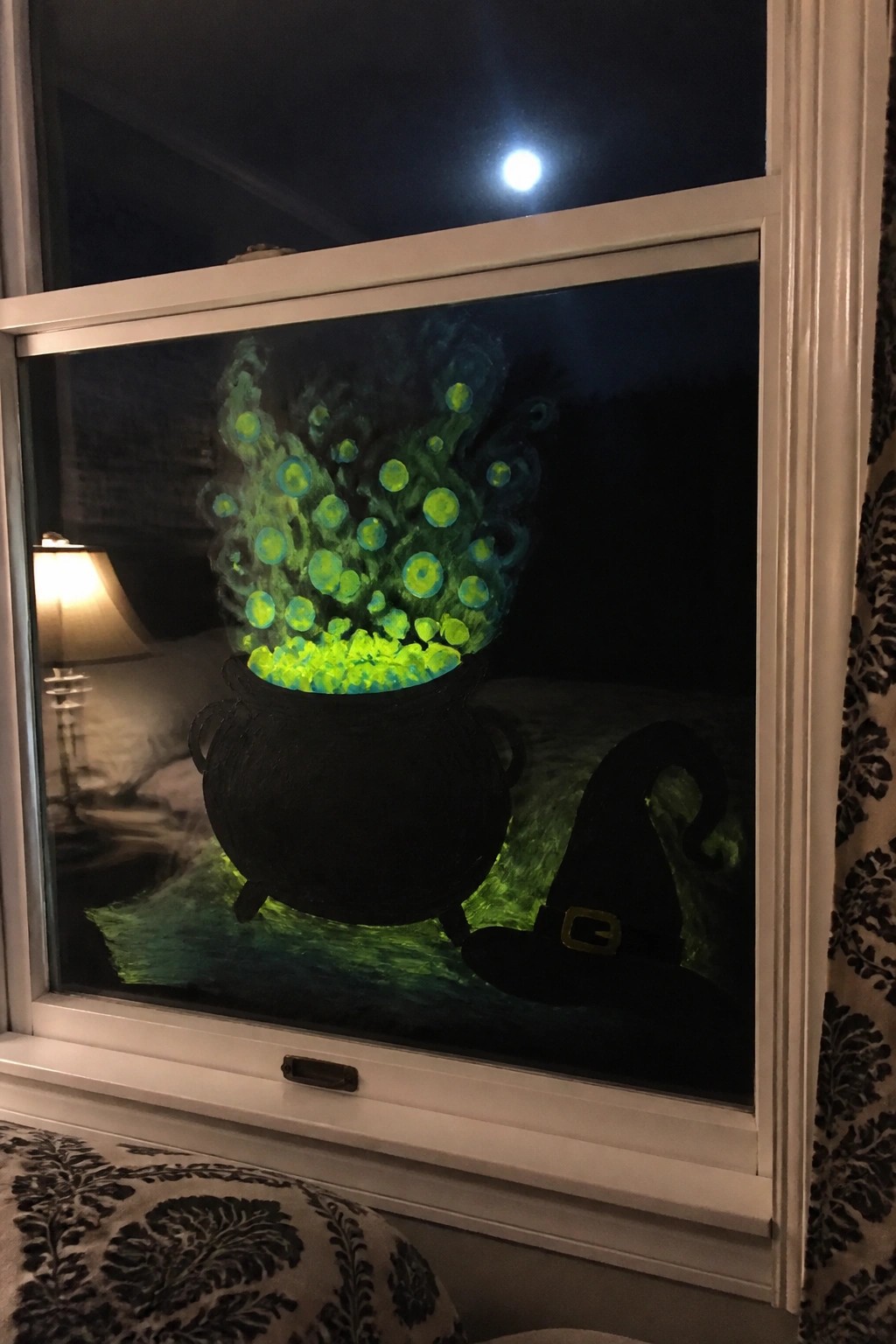 Glowing green potion bubbles from black cauldron beside witch hat on window at night.