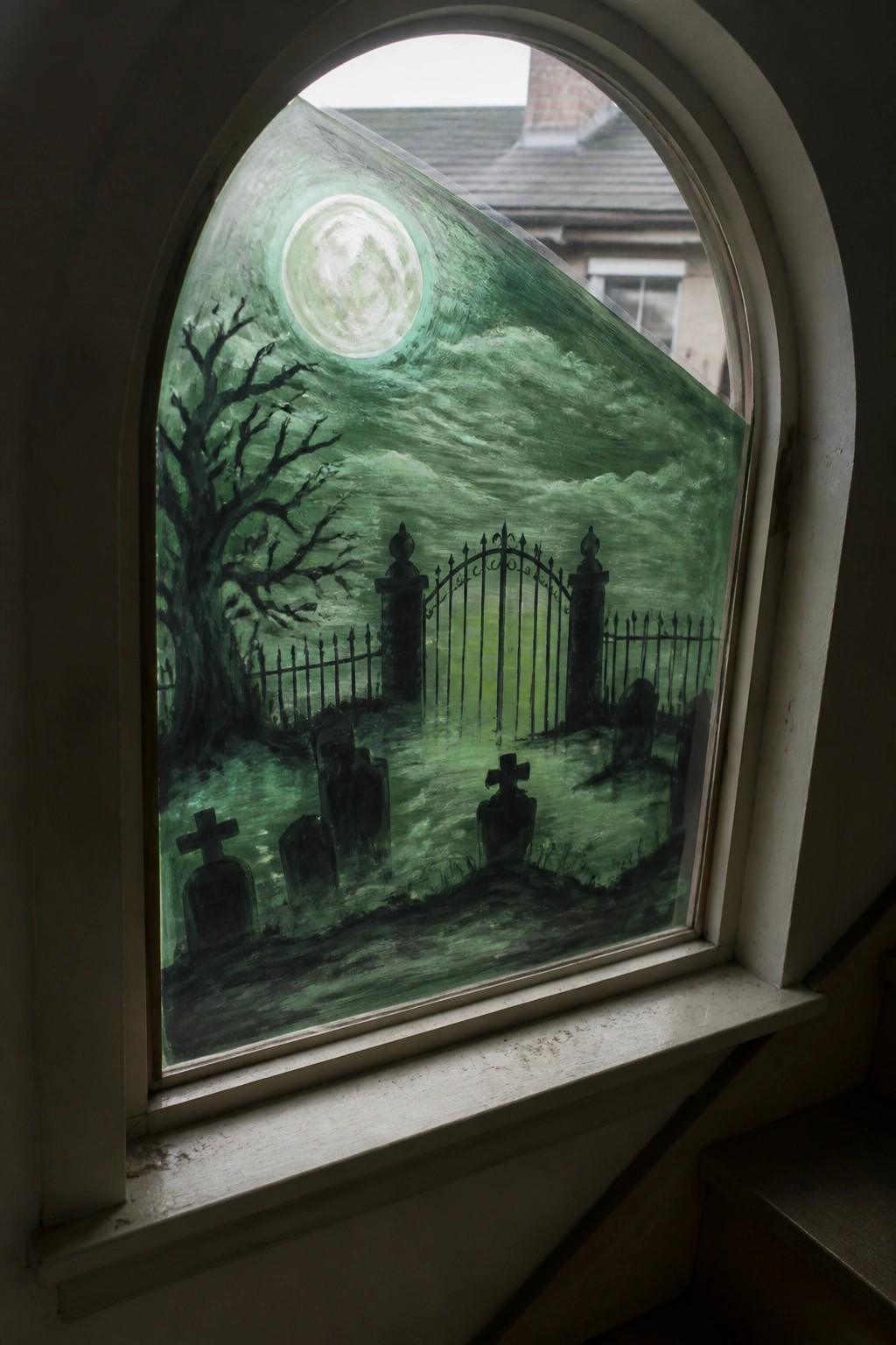 Arched window with painted green graveyard scene, full moon, gate, and tombstones.