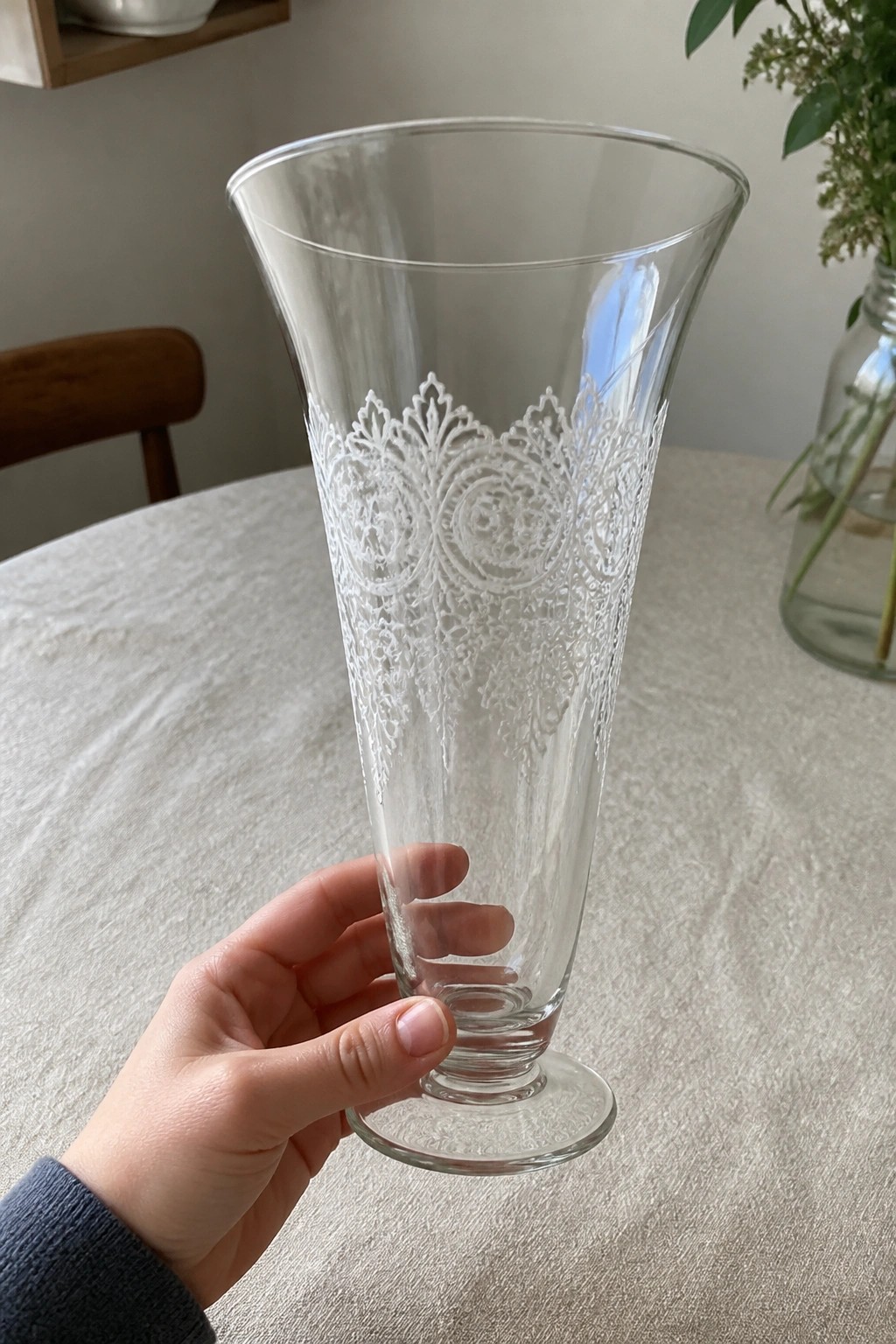 Hand holding tall clear glass vase with intricate white lace etching on table