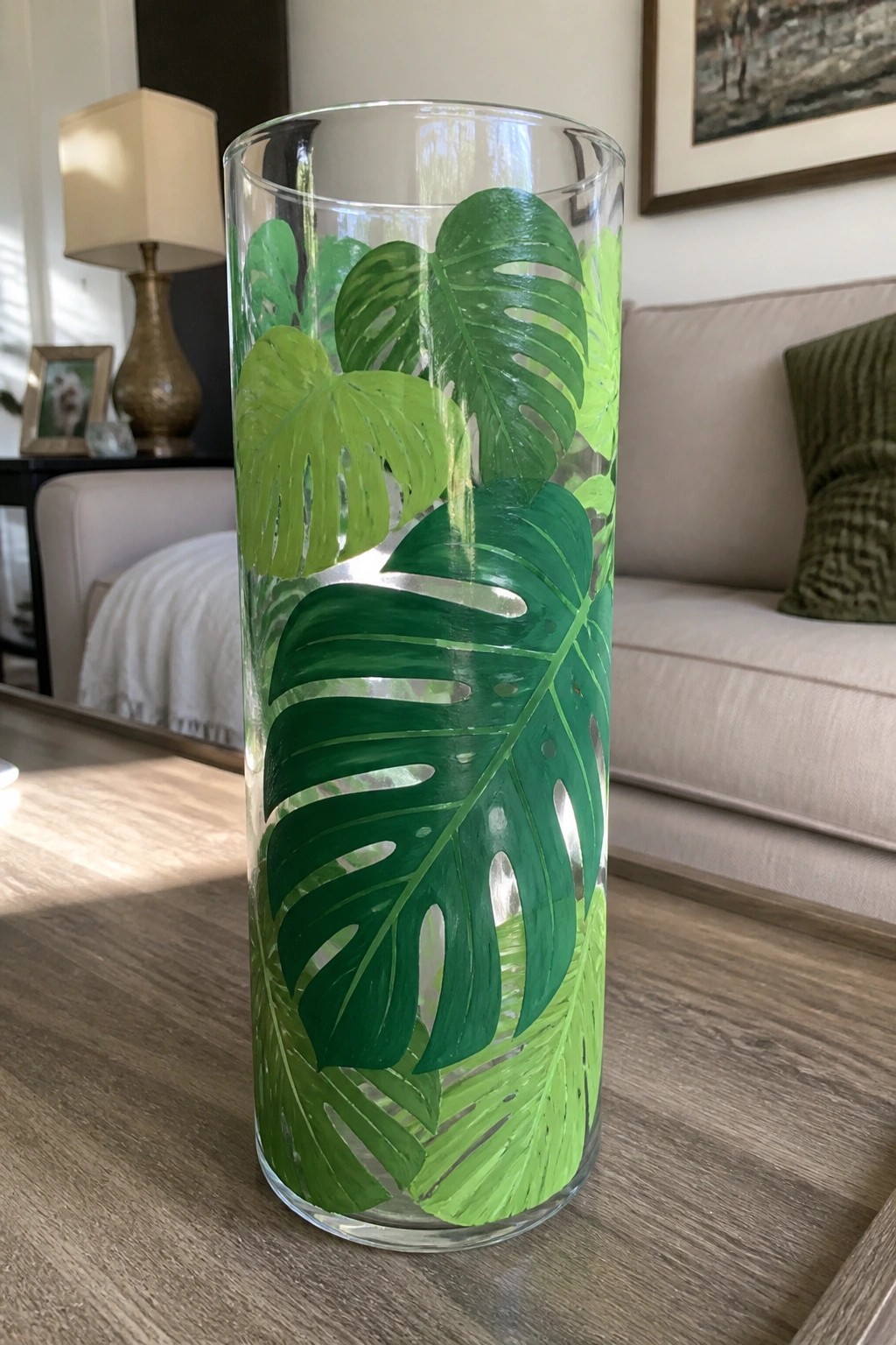 Tall glass vase hand-painted with vibrant green monstera leaves on wooden table