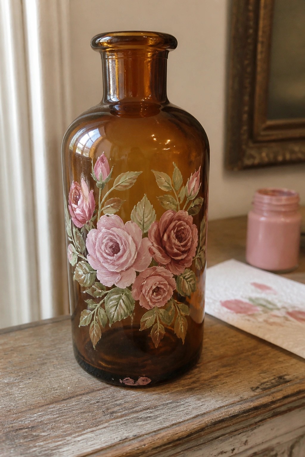 An amber glass bottle features hand-painted pink roses and green leaves on a wooden table with a pink paint jar nearby.