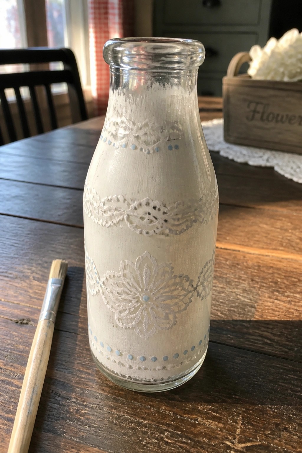 Painted glass bottle with white lace patterns, blue dots, and paintbrush on wooden table