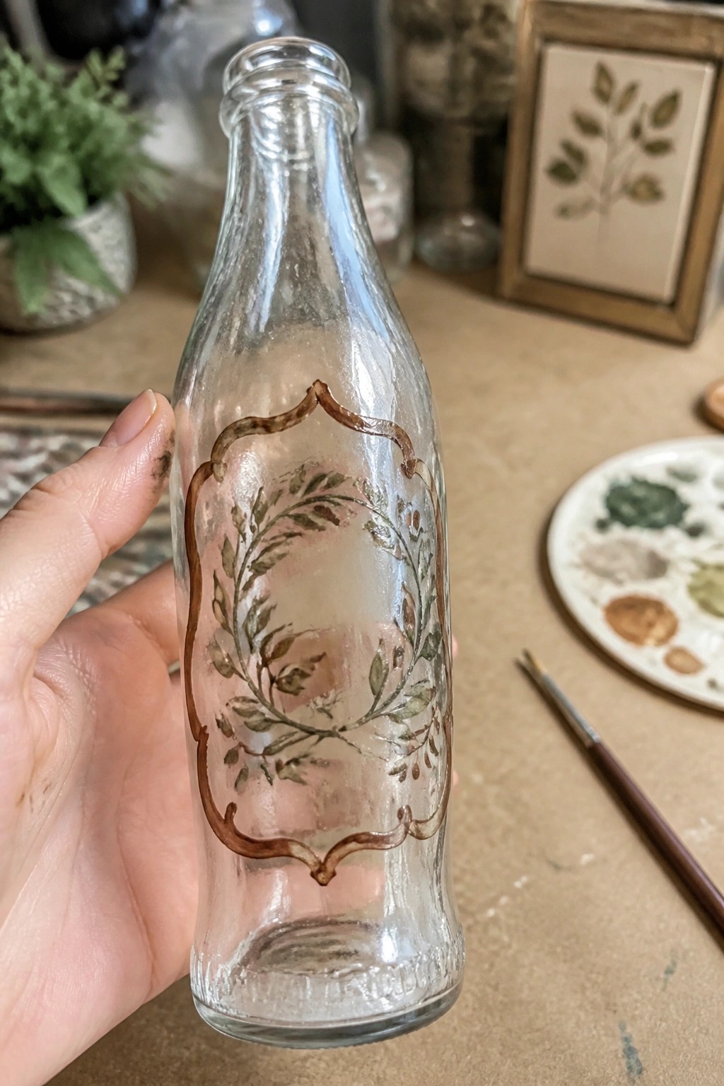Hand holding glass bottle with green leaf wreath in brown ornate border