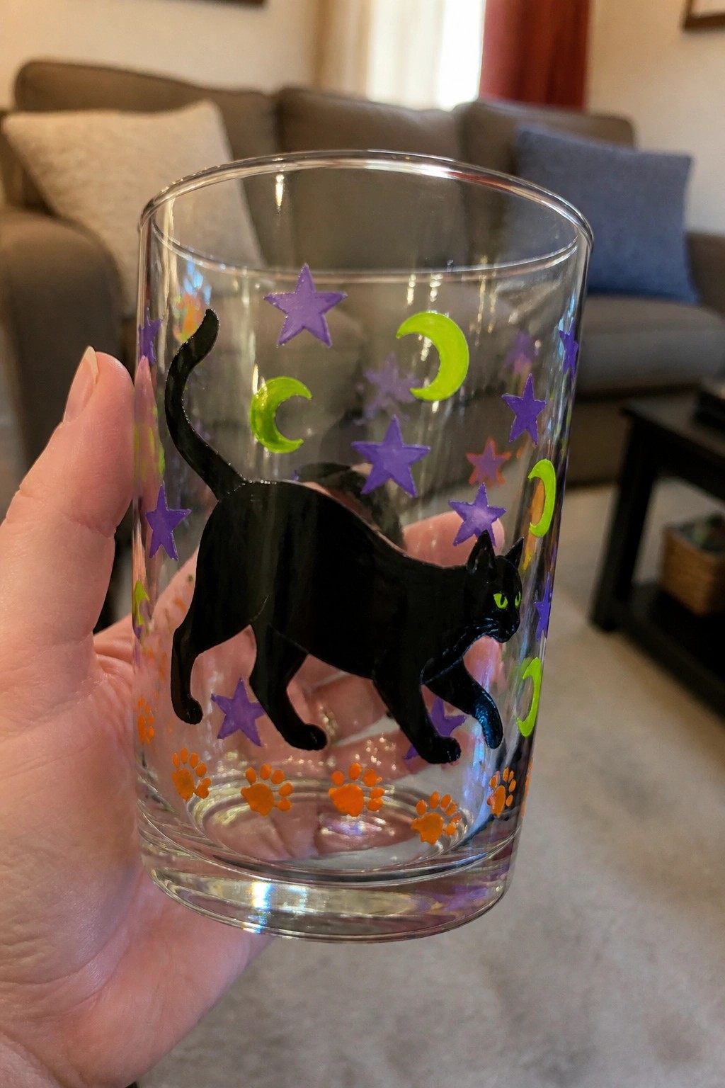 Hand holding clear glass with black cat, purple stars, green moons, orange paw prints.