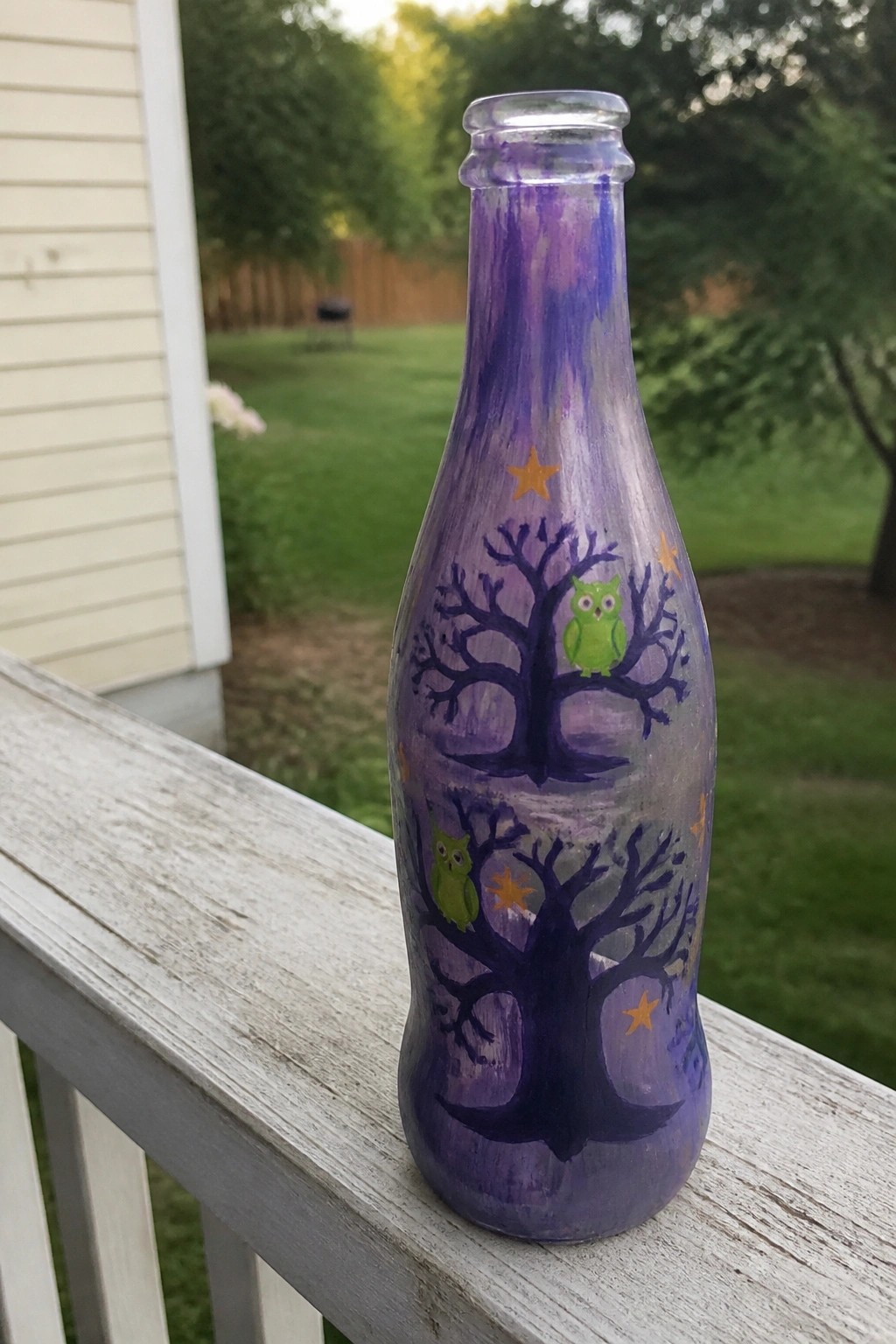 Purple painted bottle with trees, green owls, stars, and moon on wooden railing