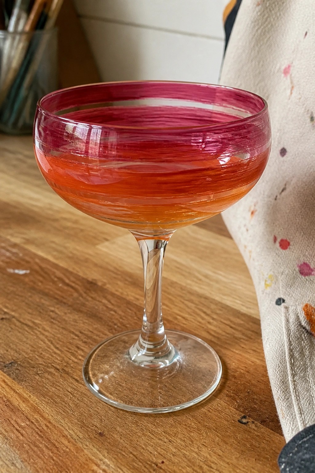 Gradient pink-orange coupe glass on wooden table beside paint-splattered cloth.