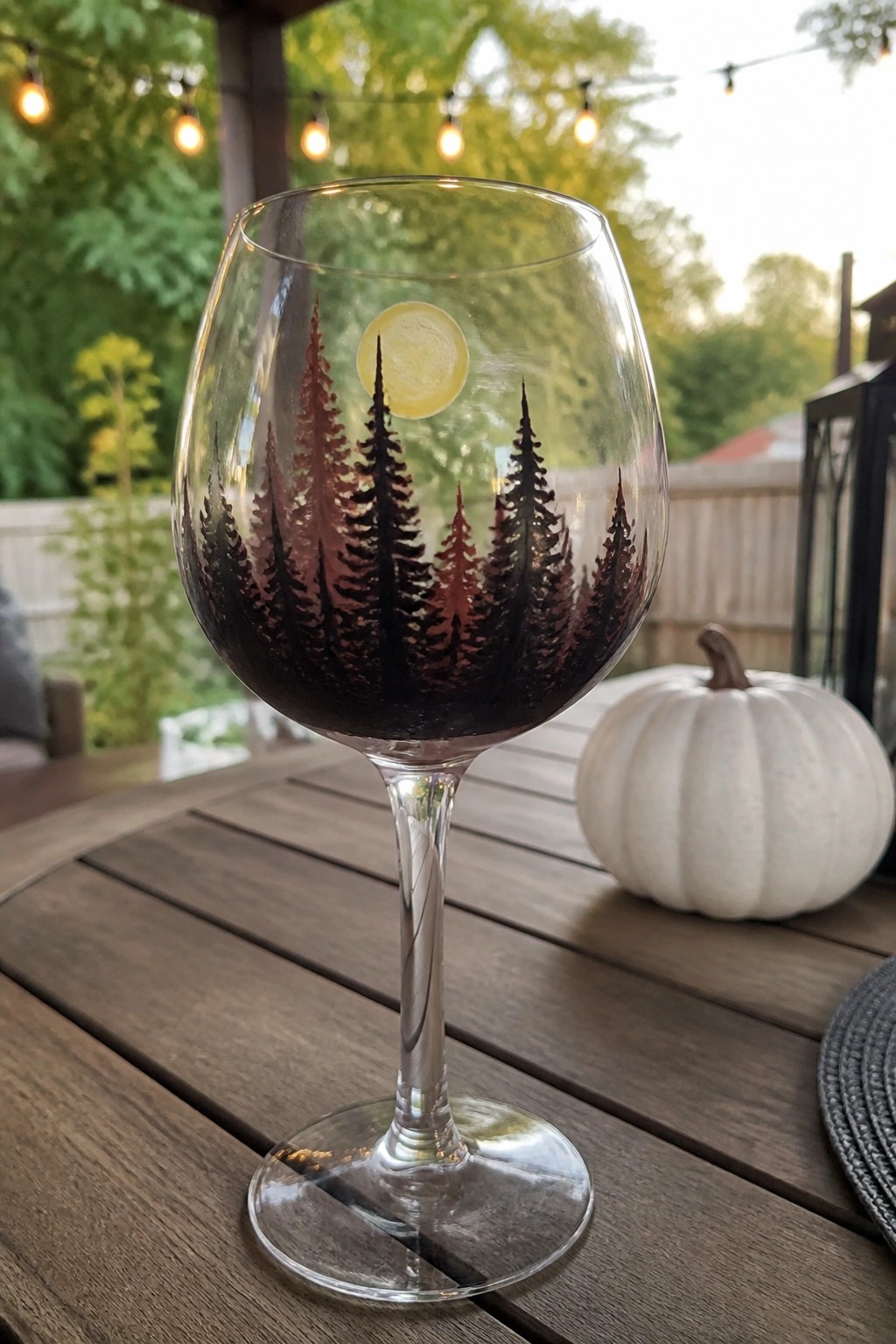 Wine glass with dark pine tree silhouettes and yellow moon, filled with red wine on table.