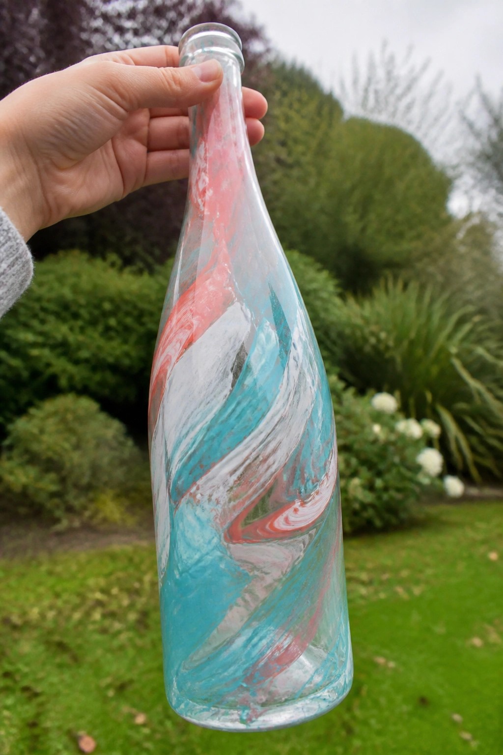 Hand holding glass bottle swirled with pink, turquoise, and white paint in garden