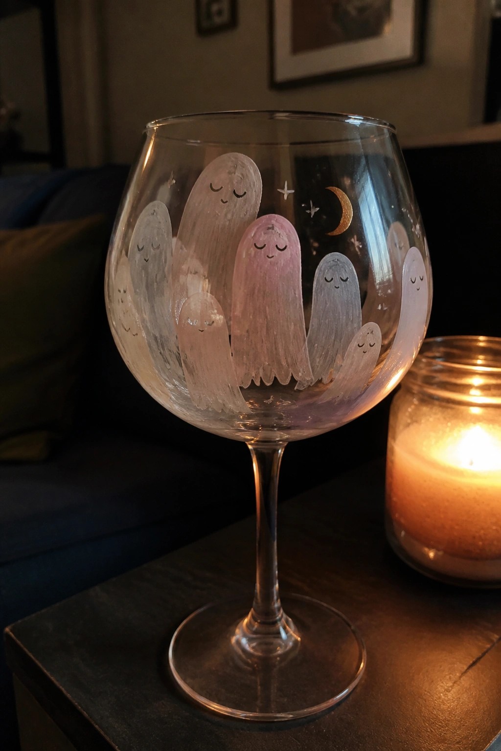 Hand-painted wine glass with pastel ghosts, moon, and stars beside lit candle.