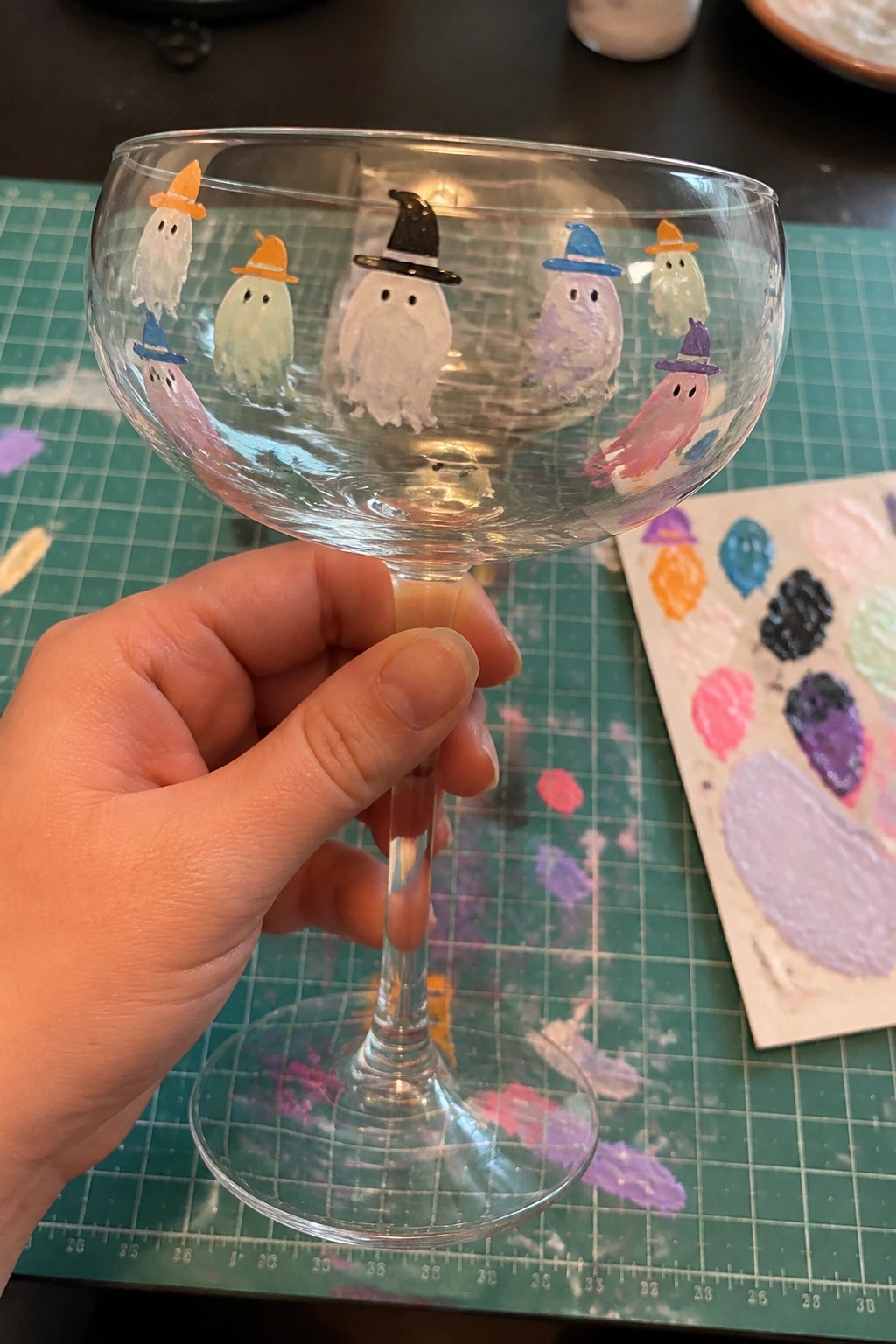 Hand holding stemmed glass with colorful painted ghosts wearing witch hats.