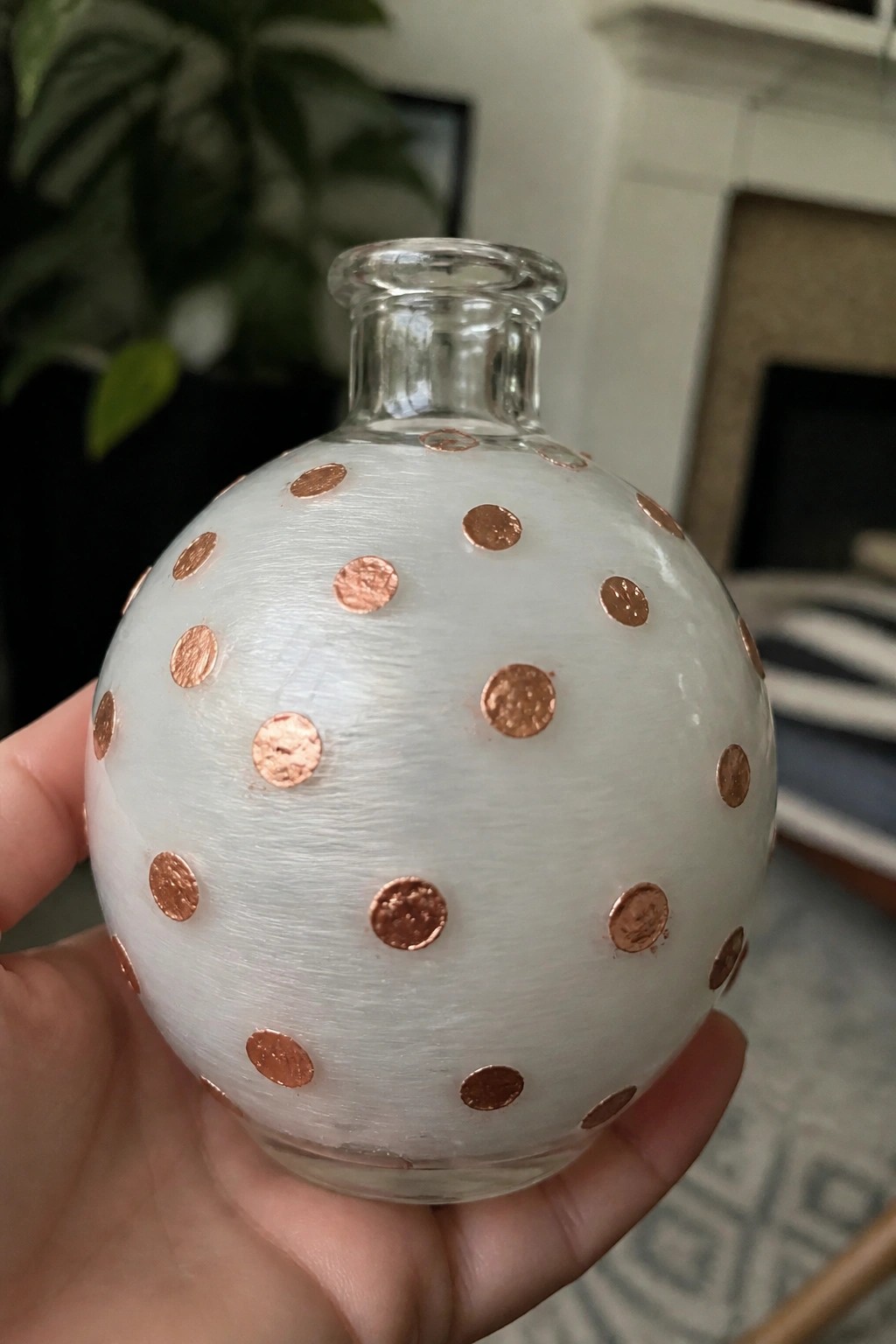 A hand holding a small round glass bottle with a frosted white body, copper metallic dots, and a clear neck.