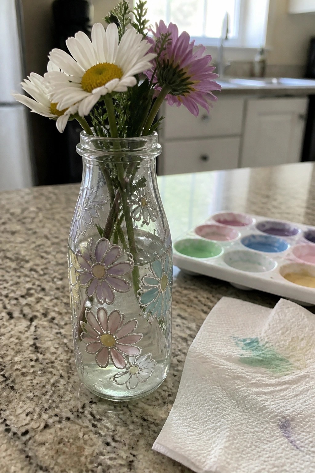 Glass vase with painted flowers holding white and purple daisies beside watercolor palette