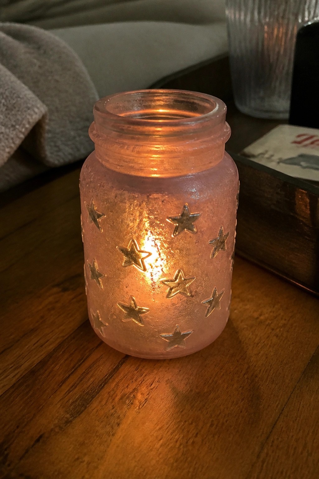 Lit candle glowing in pink frosted jar with gold stars on wooden table.