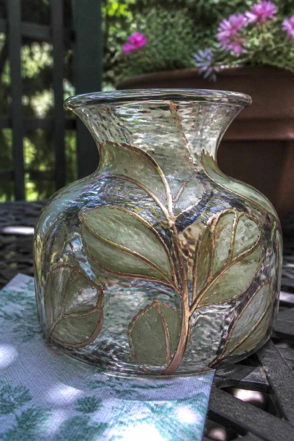 Textured glass vase with green leaves and gold stems on patterned cloth.