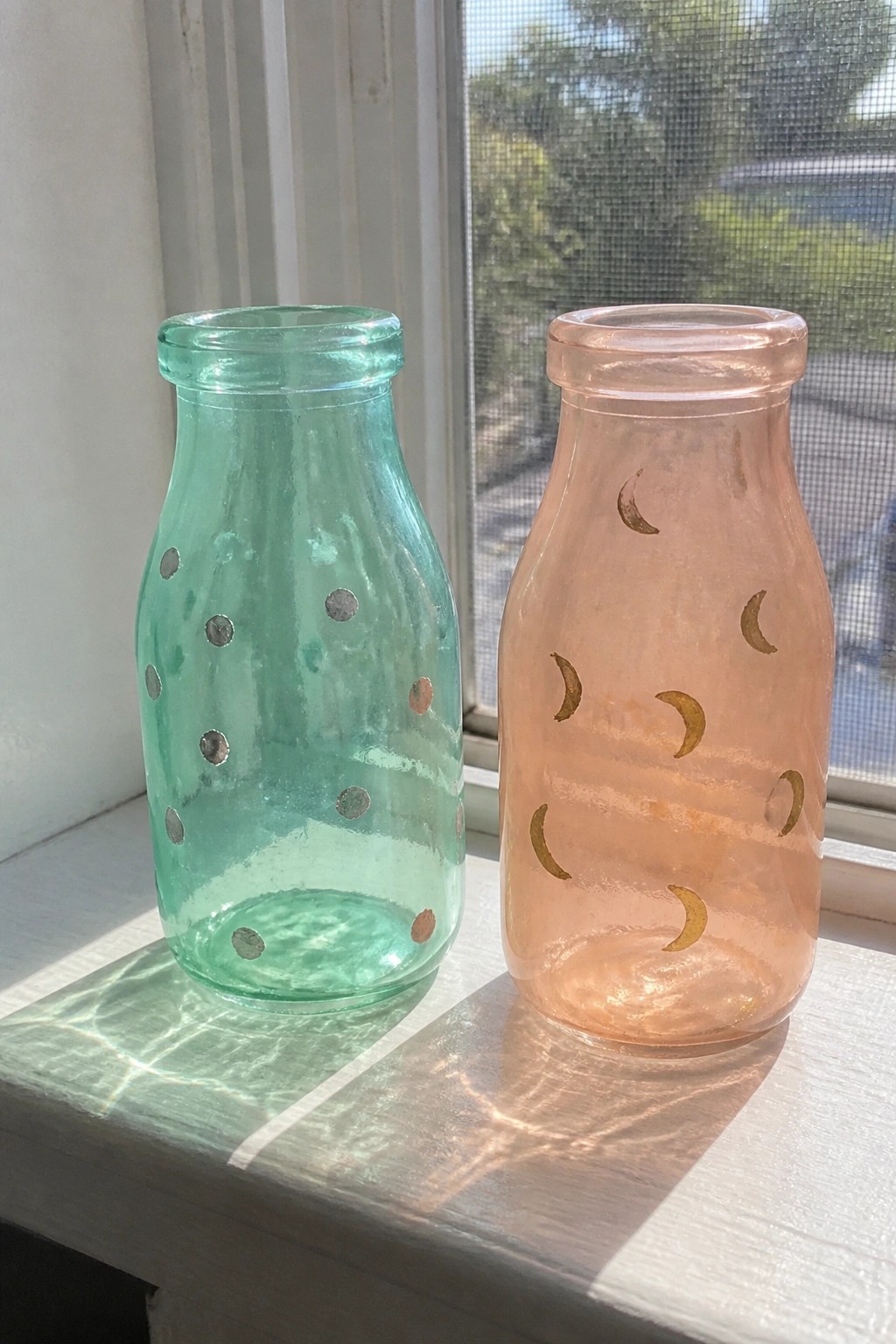 Two glass bottles with metallic dot and moon designs sit on a white windowsill.