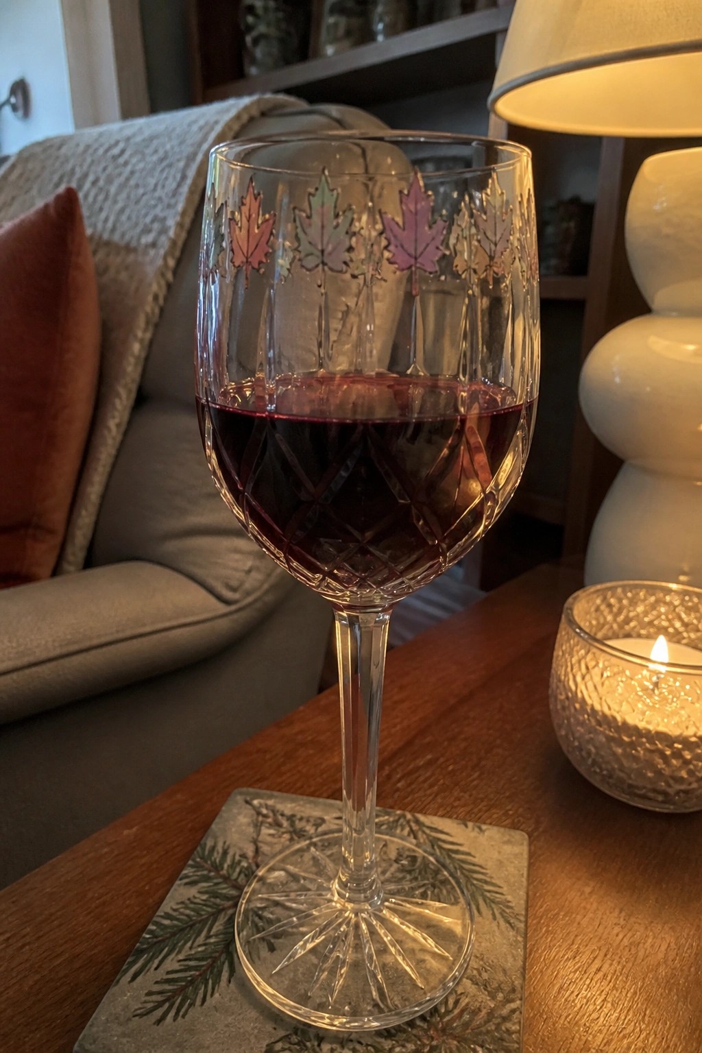 Crystal wine glass etched with colorful maple leaves holding red wine beside lit candle