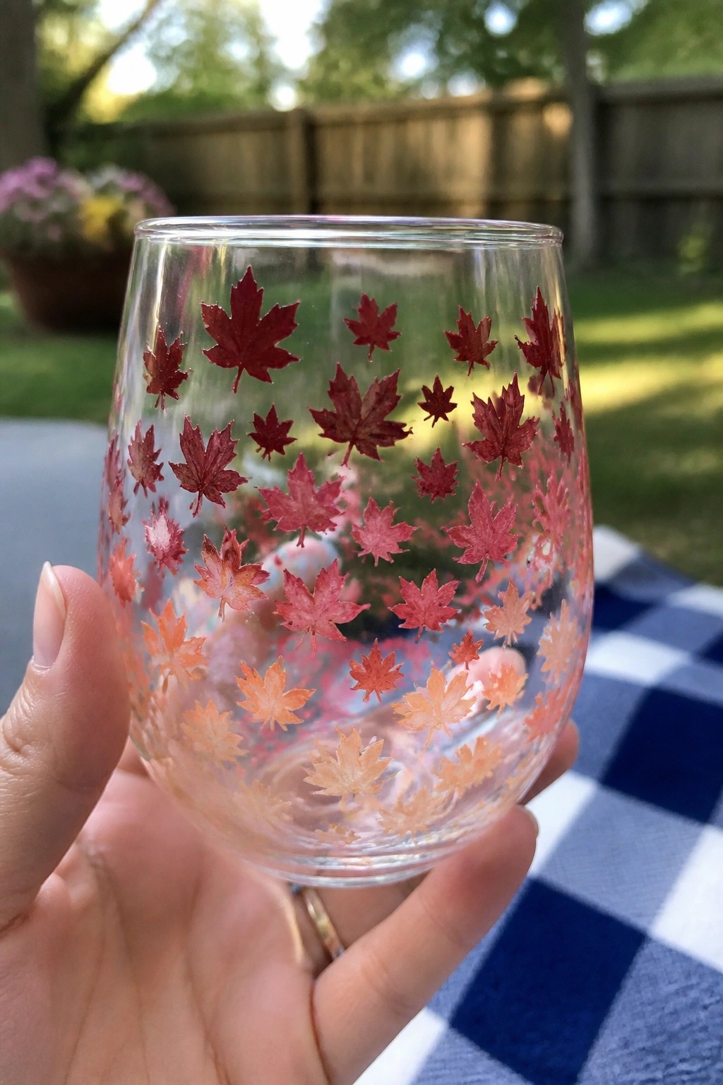 Hand holding clear glass with red-to-orange maple leaf decals on checkered blanket
