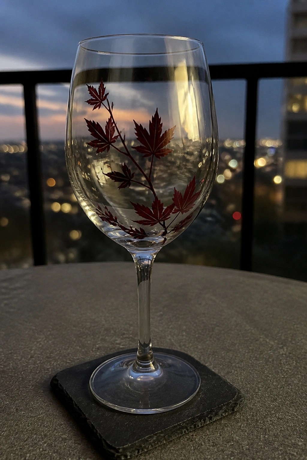 Wine glass etched with red maple leaves on dark coaster overlooking dusk city lights