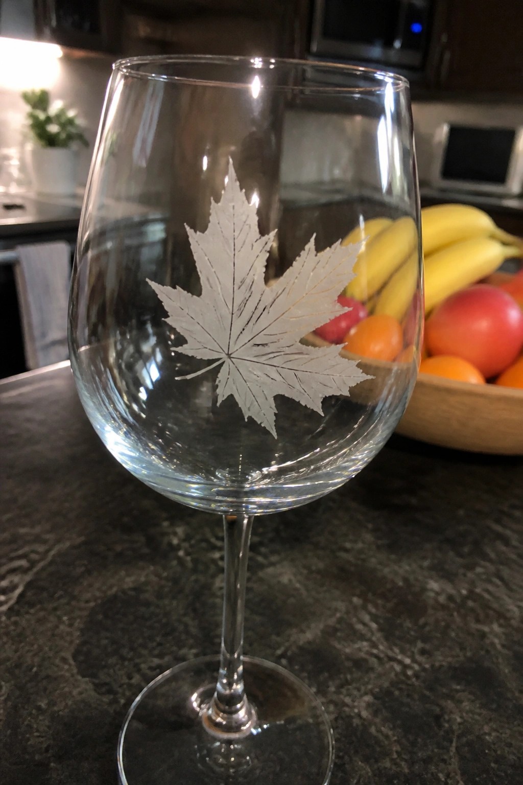 Wine glass with etched maple leaf on dark countertop beside fruit bowl