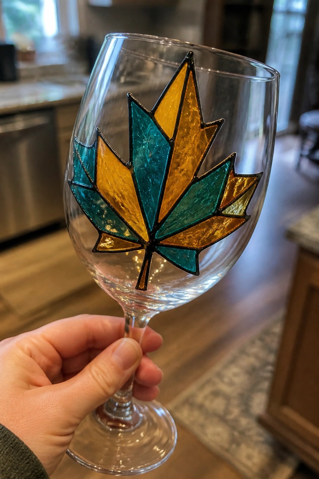 Hand holding wine glass with teal and amber stained-glass maple leaf design.