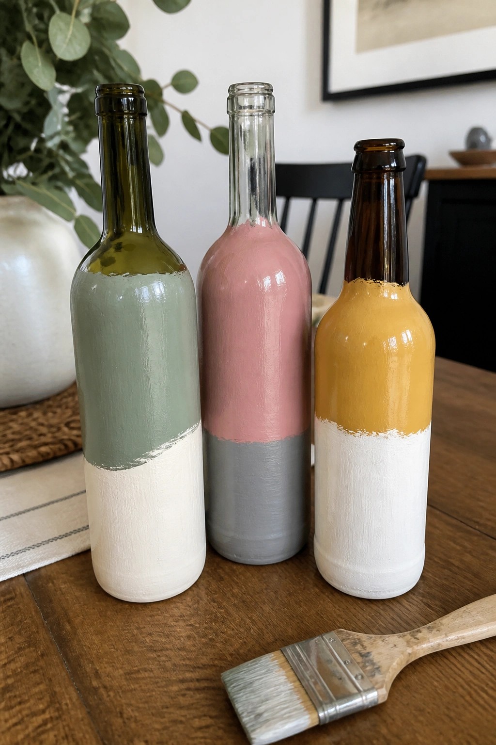 Three two-toned painted bottles in green, pink, and yellow with a paintbrush on wood table.