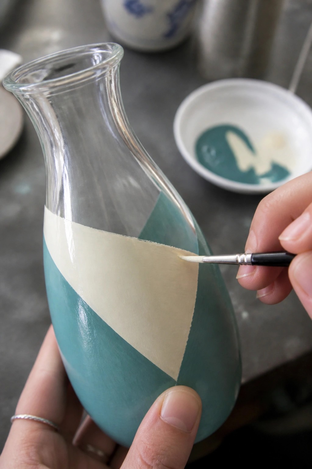 Hands painting teal geometric stripes on glass vase using brush and tape.