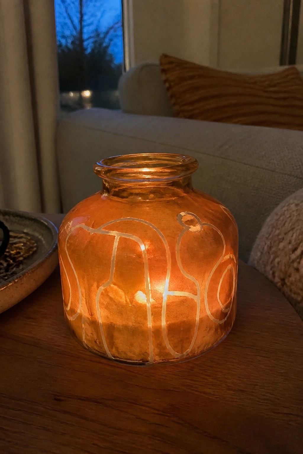 Lit candle glowing inside orange glass jar etched with white pumpkins on wooden table