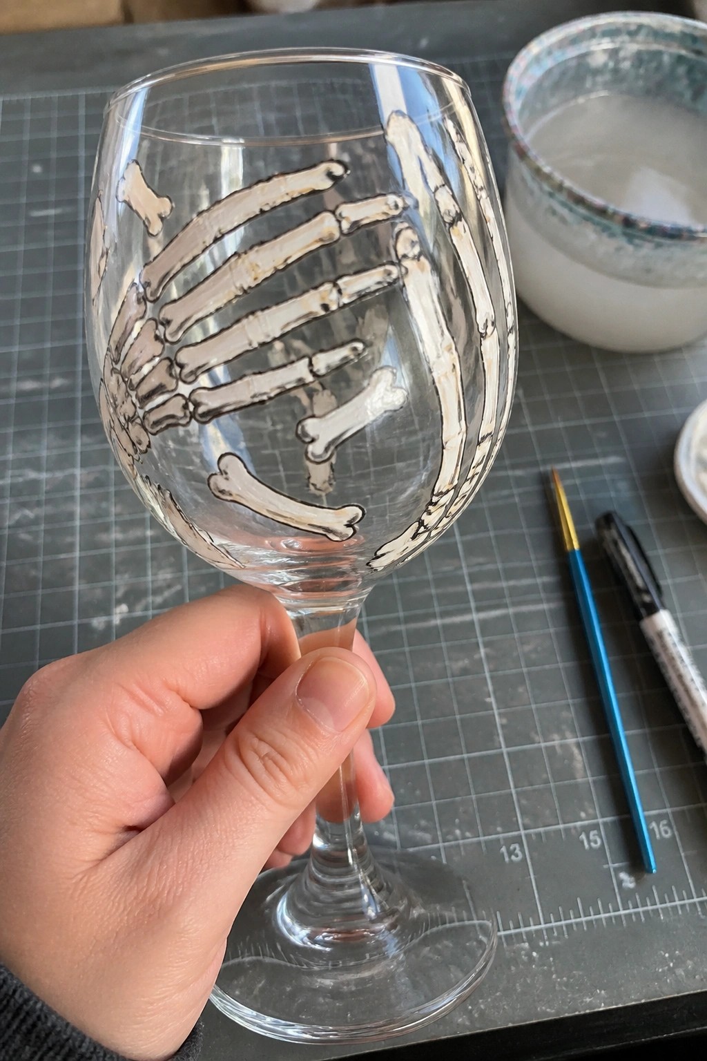 Hand holding wine glass with white skeletal hand painting amid art supplies on grid mat.
