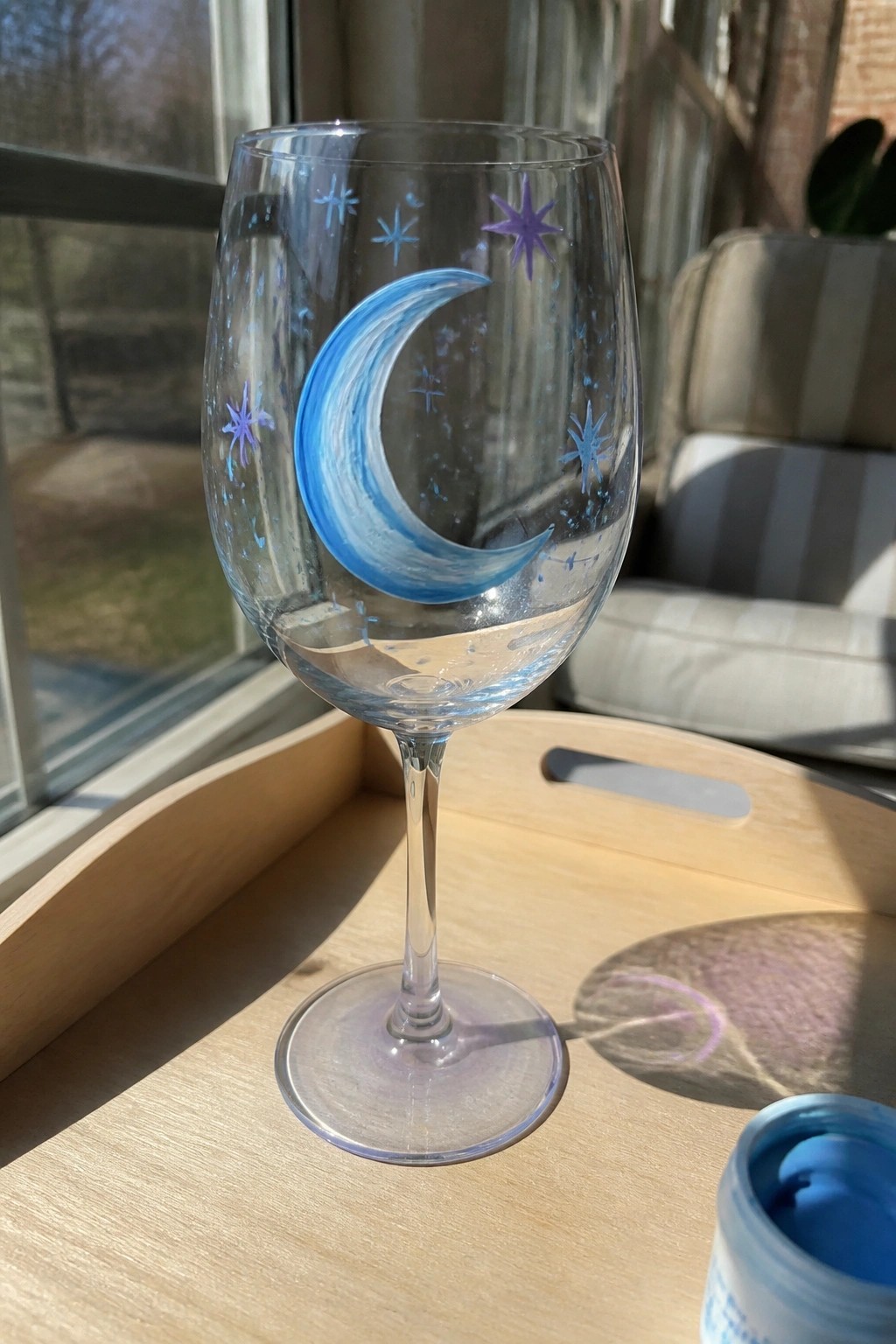 Hand-painted wine glass with blue crescent moon and stars on wooden tray in sunlight