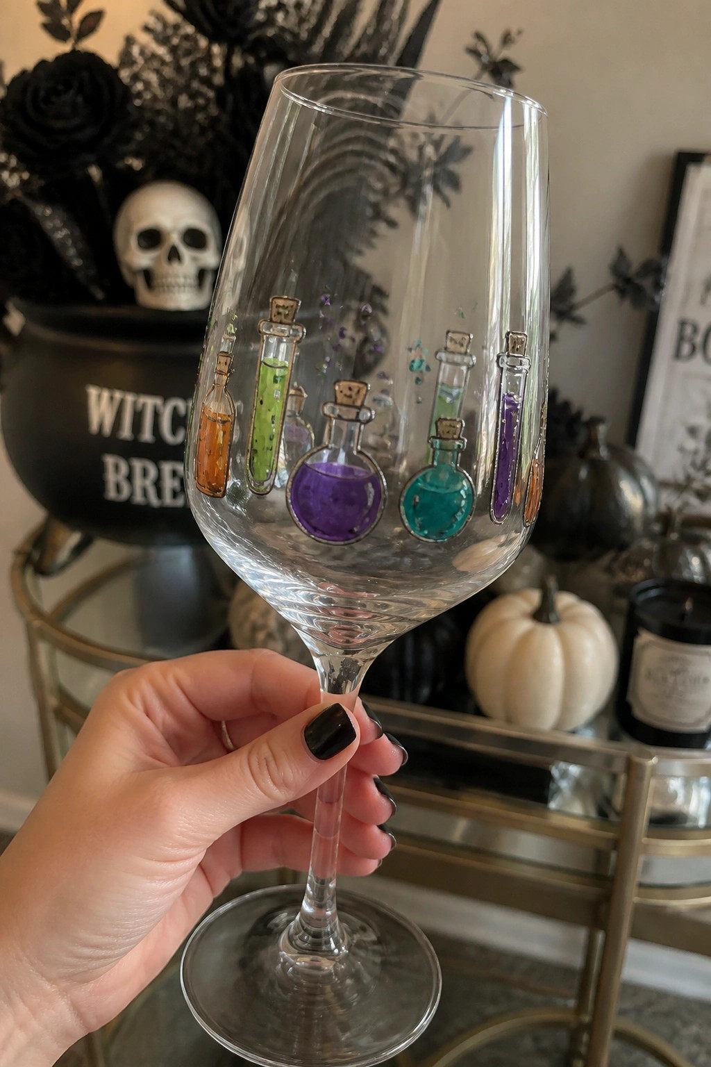 Hand holding wine glass with colorful potion bottle decals, Halloween cauldron and skull backdrop.