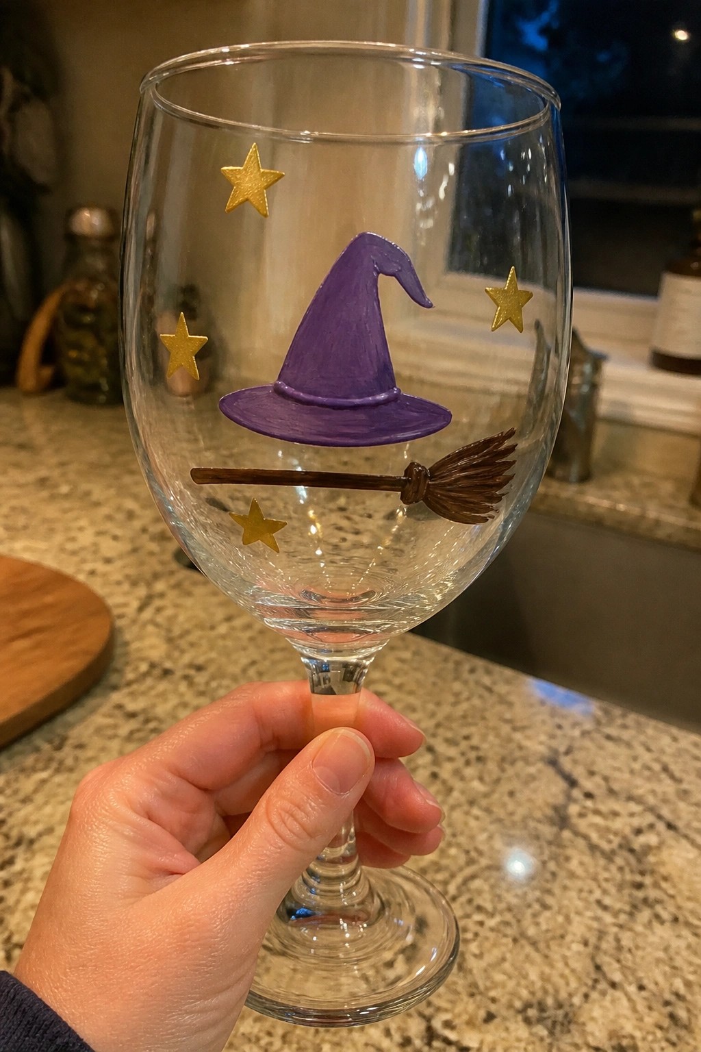 Hand holding wine glass painted with purple witch hat, broom, and gold stars