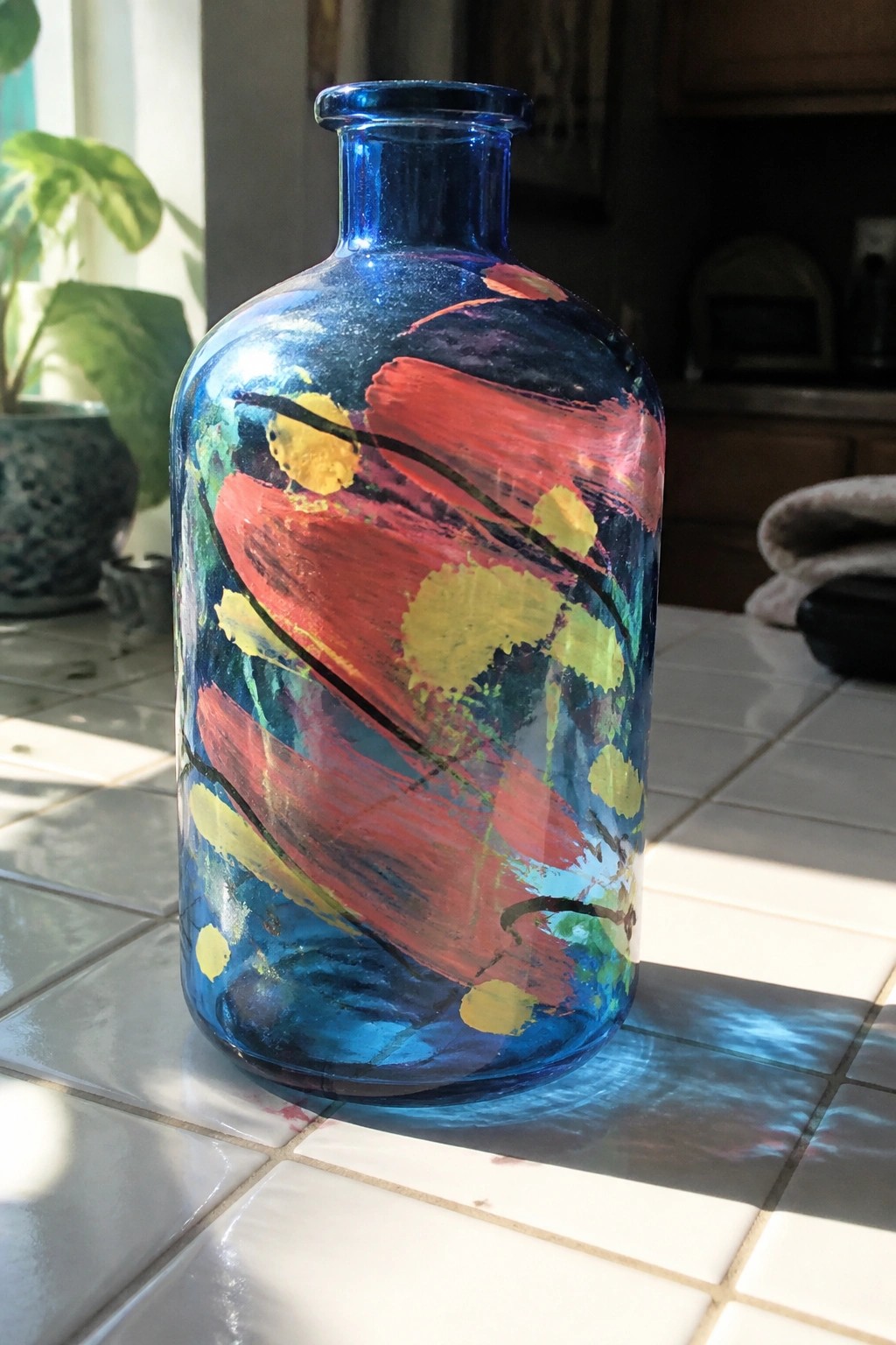 Colorful abstract-painted blue glass bottle on a sunlit white tiled countertop.