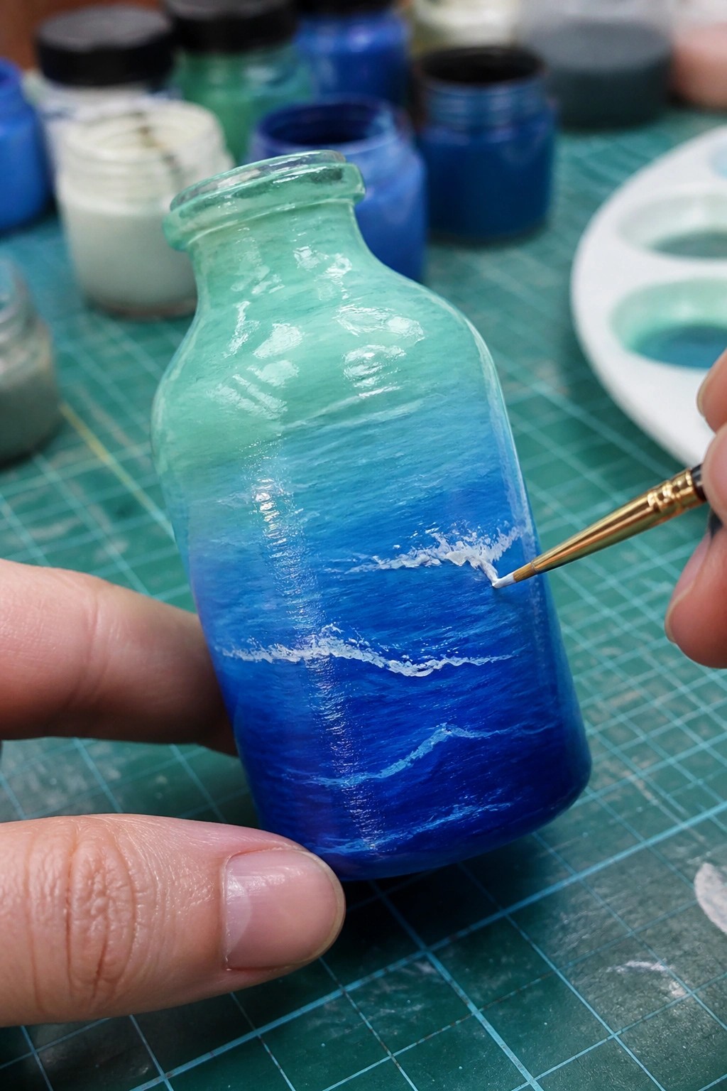 Hand painting white waves on gradient blue ocean-themed glass bottle with fine brush.