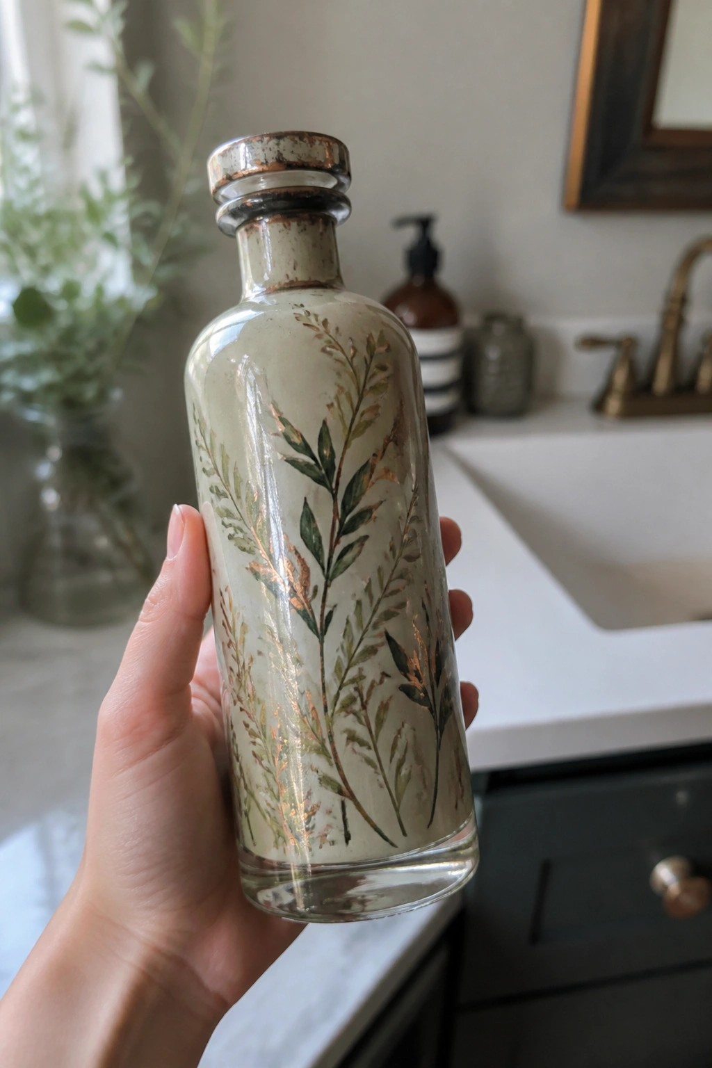 Hand holding glass bottle with green leaves and copper branch designs in bathroom