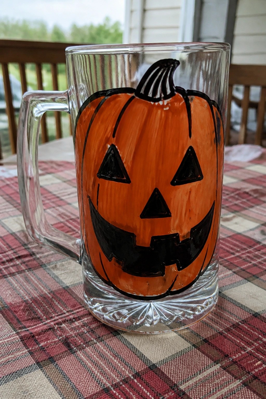 A clear glass mug with an orange and black painted jack-o'-lantern face sits on a red plaid tablecloth.